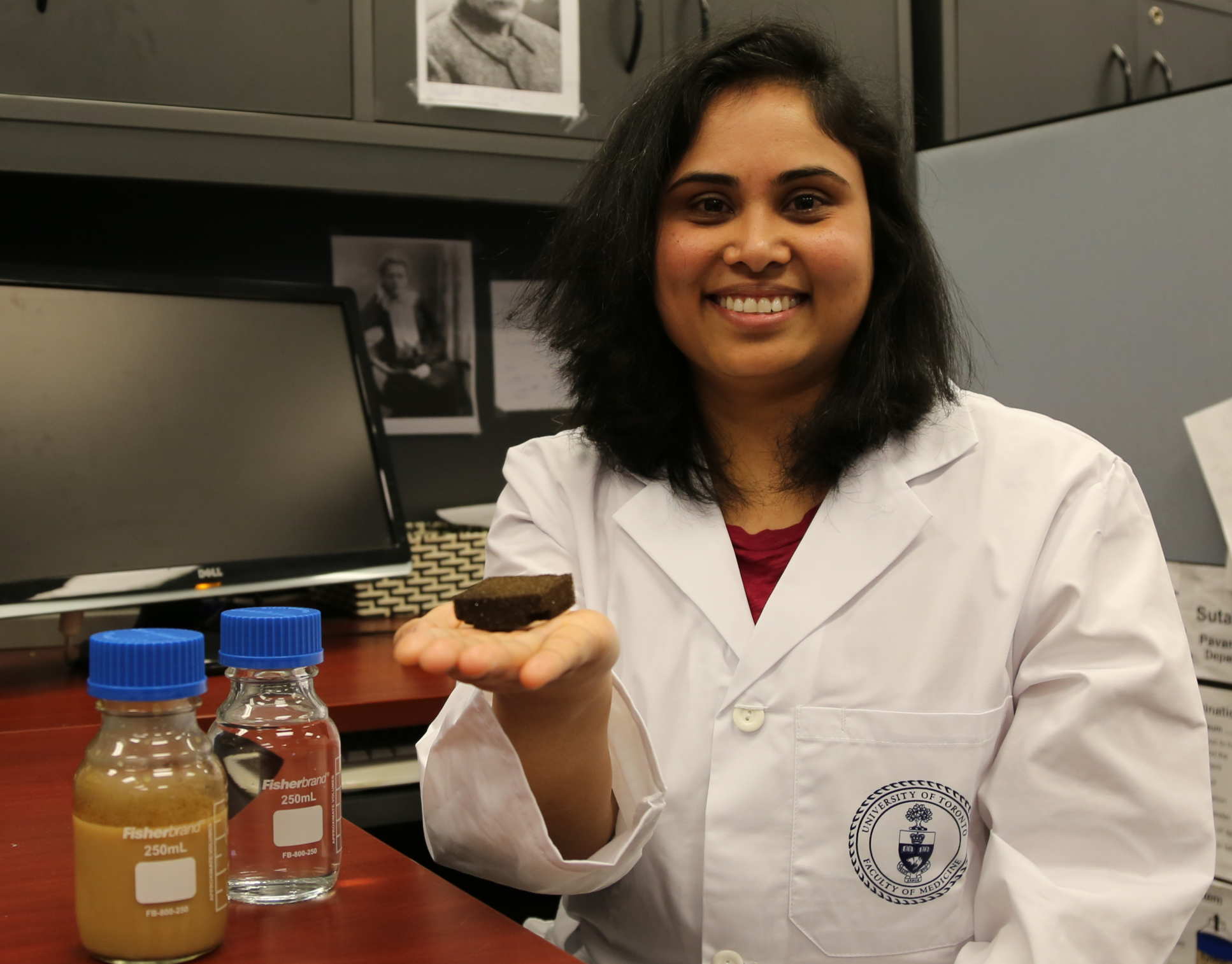 Dr Pavani Cherukupally with the a bottle of brown oil-contaminated water, a bottle of clean and clear water, and the new sponge. She is sitting at her lab bench and smiling at the camera, sponge in hand.
