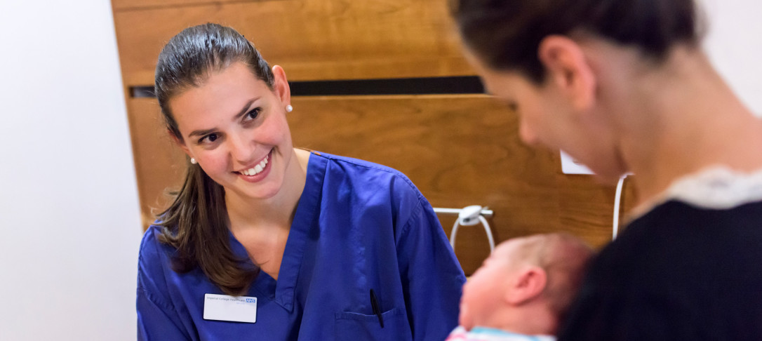A nurse in blue scrubs smiling at a mother and newborn baby