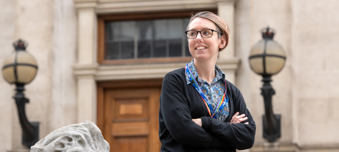 Photo of Caz Ulley, a woman with short hair wearing glasses, dressed in a black cardigan over a blue shirt, wearing a rainbow lanyard, with the Imperial Queen's Tower as a background