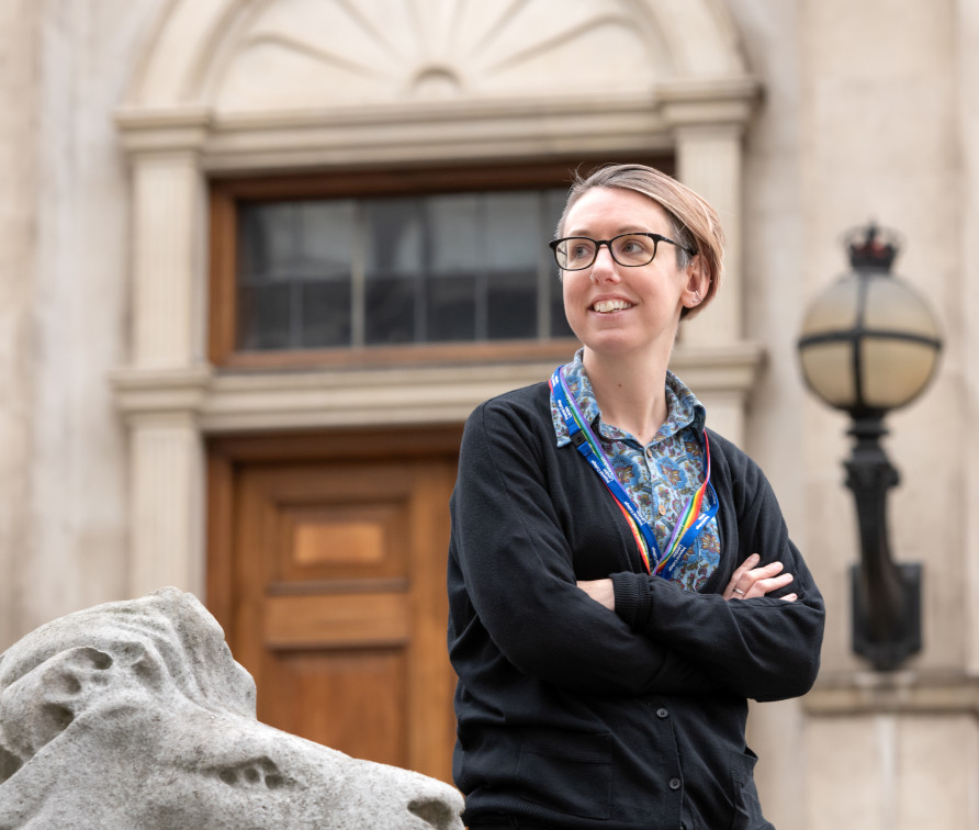 Photo of Caz Ulley, a woman with short hair wearing glasses, dressed in a black cardigan over a blue shirt, wearing a rainbow lanyard, with the Imperial Queen's Tower as a background