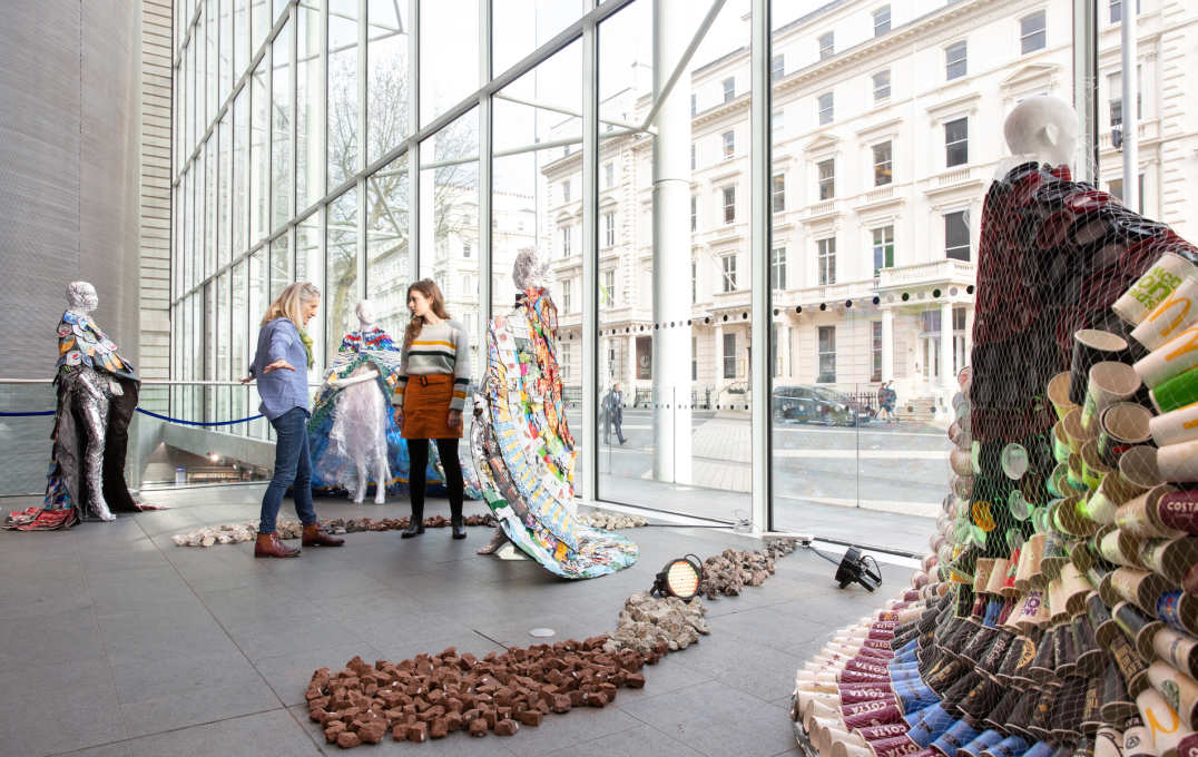 Two women stand in an airy atrium amongst mannequins, as people and traffic pass by in the street outside the window