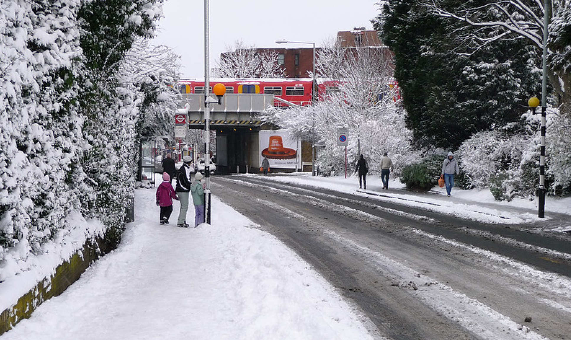 A wet, snowy roadside in an English town, woman and two kids wrapped in hats, coats and boots wait at a bus stop. People trudge along icy road opposite, a train passes on a brige over the empty road