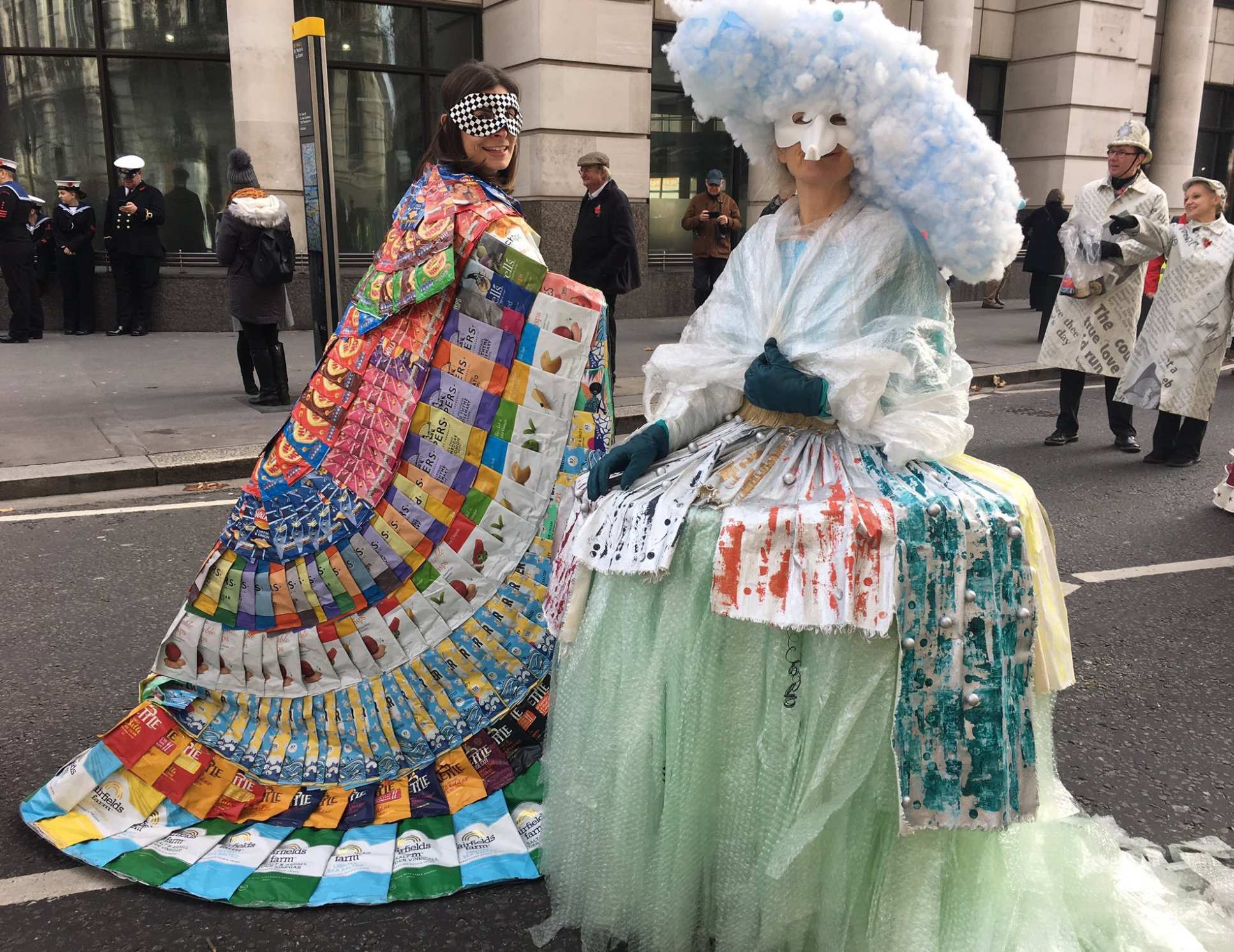 Two people wear cloaks made of trash. Left: crisp packets. Right: discarded plastics including bubble wrap and polystyrene