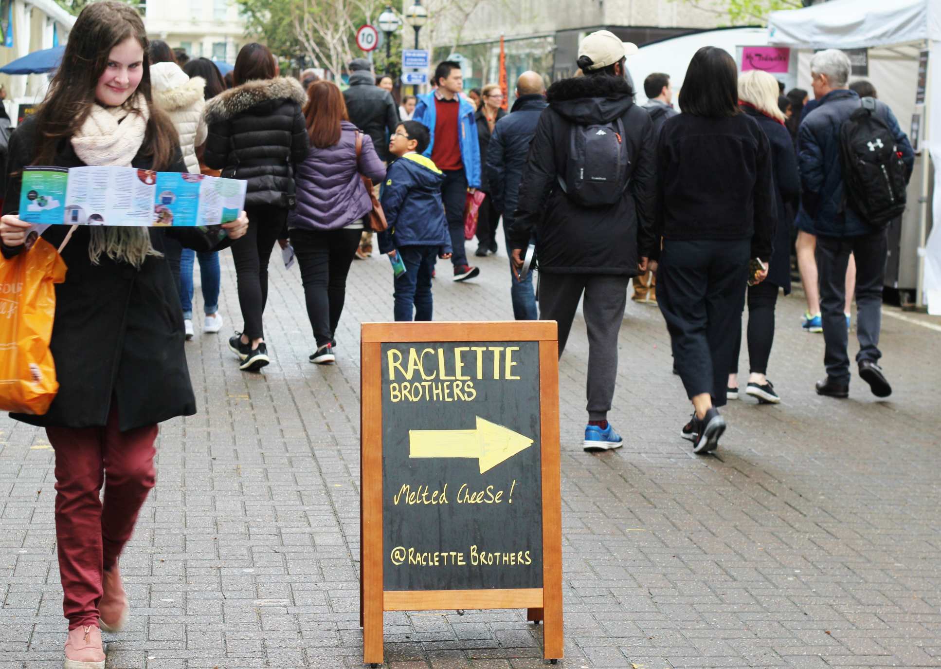 A street of food stalls with a sign offering raclette
