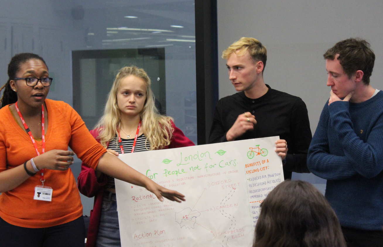 Four students present a colourful poster in front of a room of their peers