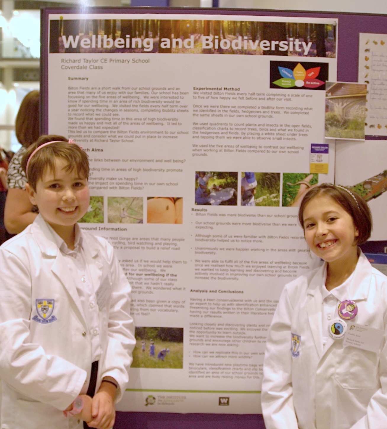 Photo of two children in lab coats presenting their work in front of a poster titled "Wellbeing and Biodiversity"