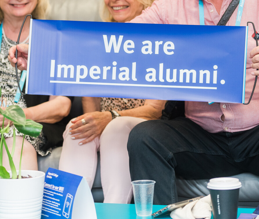 A group of senior alumni, faces not visible, at an event. One holds up a purple pull-out banner that reads 'We are Imperial alumni.' 