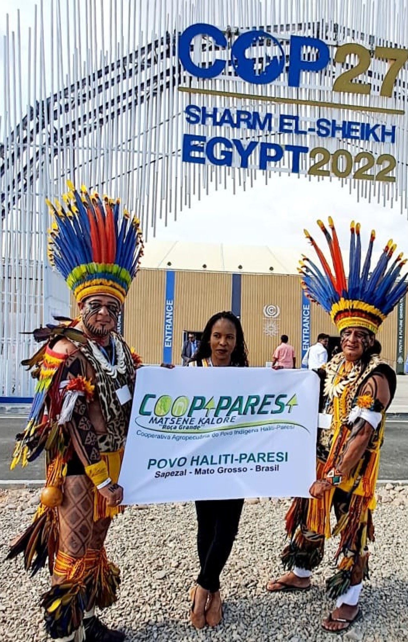 Three people stand outside a conference venue, two are wearing traditional outfits of Indigenous people of Brazil, they hold up a sign in Portuguese