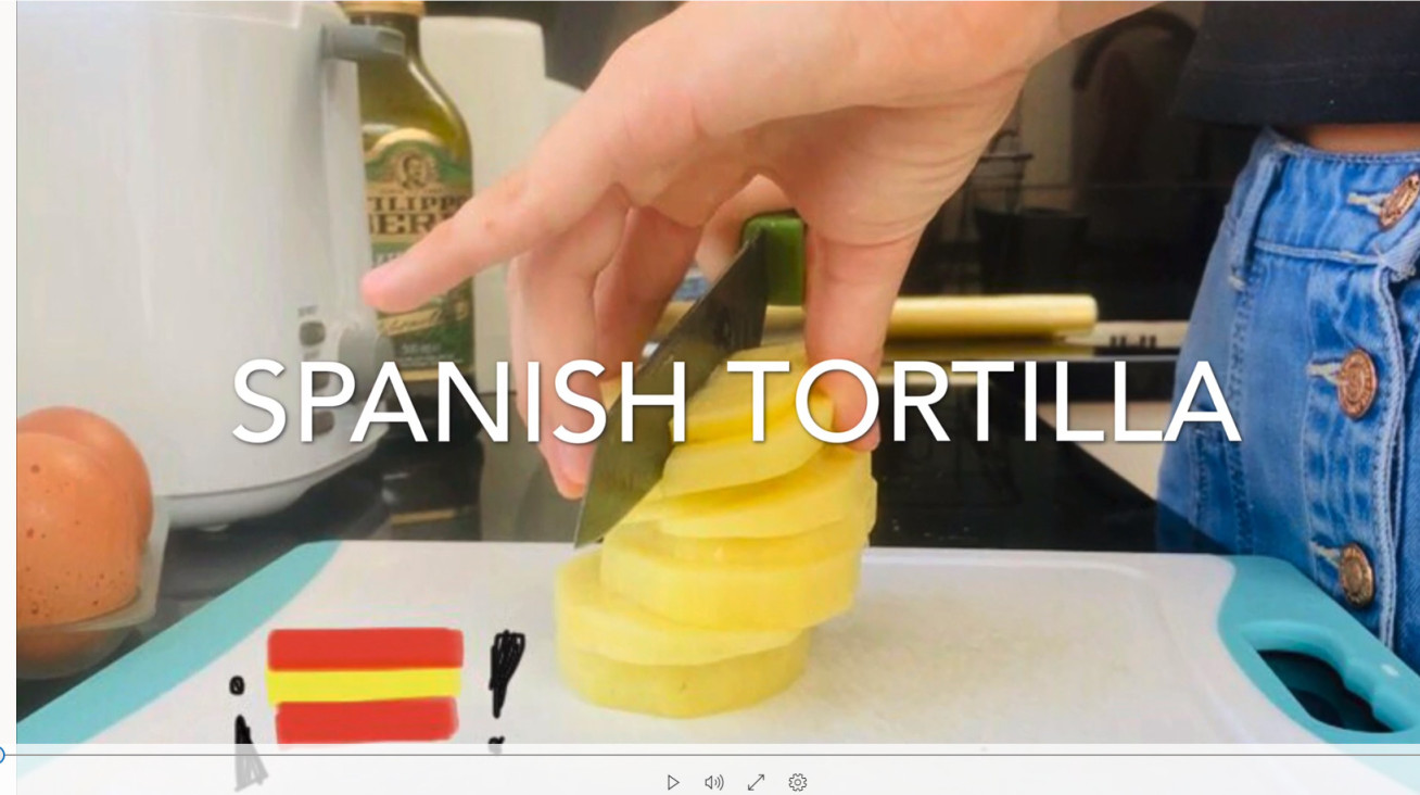 Still from a video - text: Spanish Tortilla on top of a hand chopping a stack of potatoes