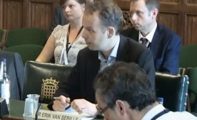 Man sits at a desk with a name plate in front of him next to a house of commons crest