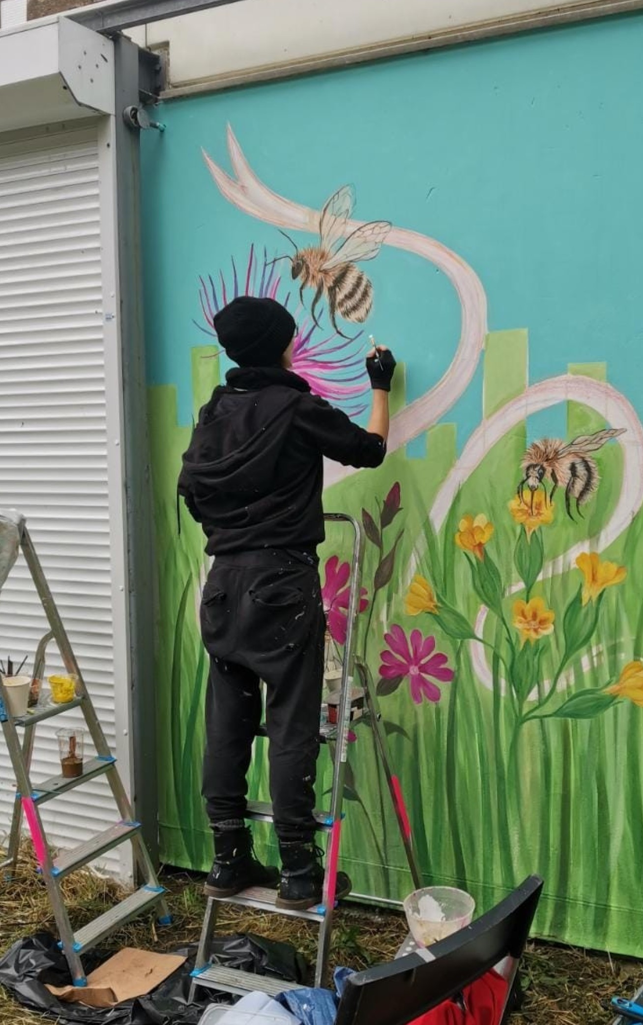 Photo showing East London artist Michelle Meola painting the wall mural in Tower Hamlets Cemetary Park, she is dressed in black and surrounded by paint pots