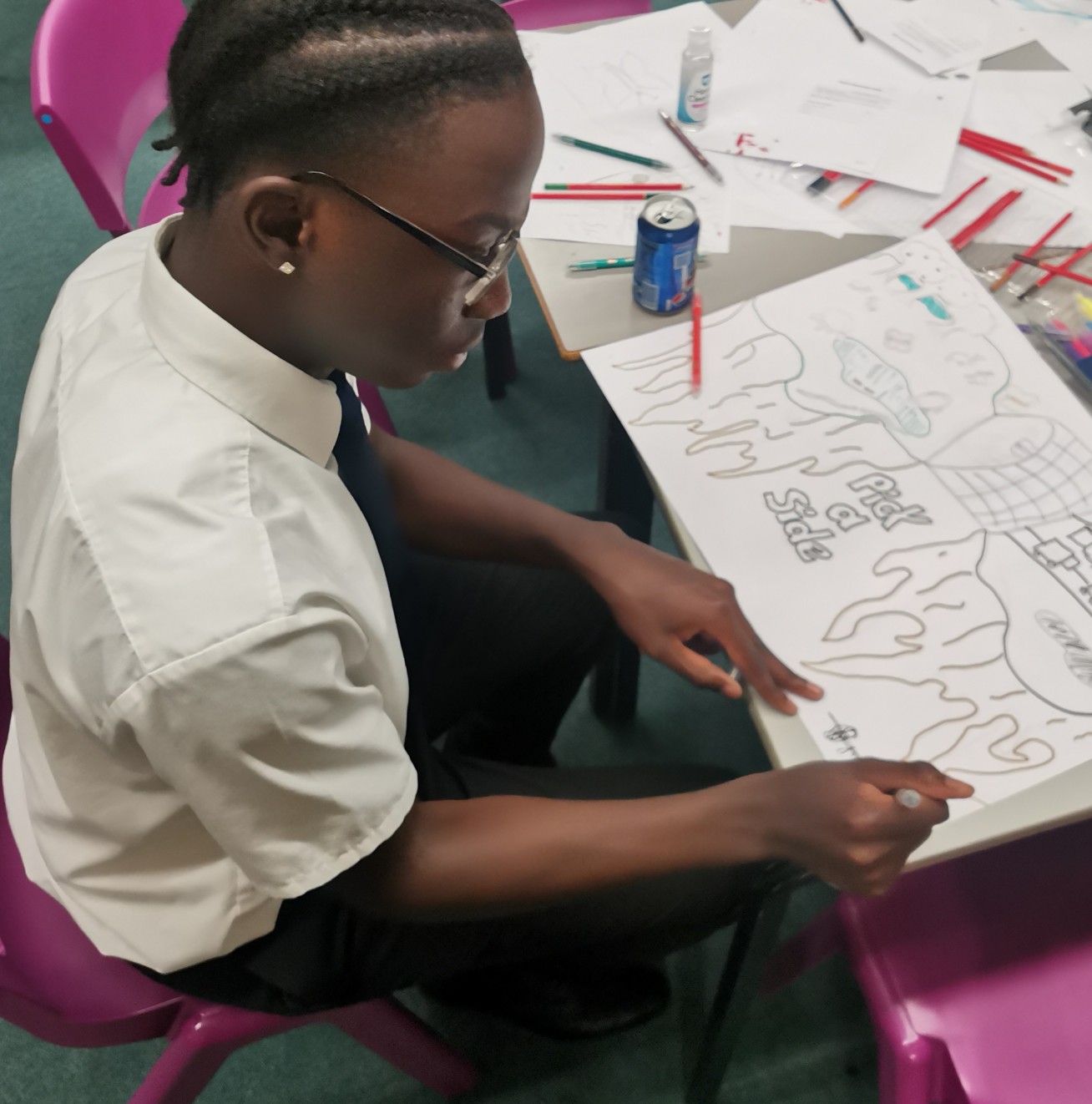 Photo shows a Black boy in school uniform sitting at a desk drawing on a large sheet of paper, it reads 'pick a side' and shows a melting glacier