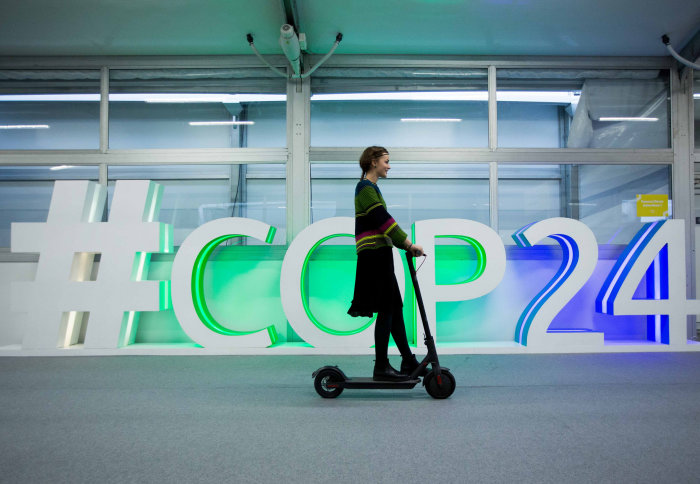 Woman rides scooter in front of white, green and blue sign reading hashtag COP24