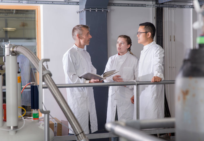 Two students in lab coats talking to a professor in an engineering lab