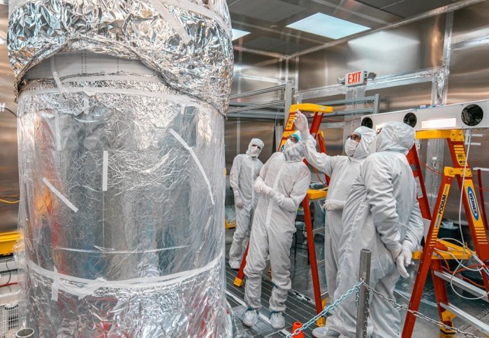 People in full white hazmat suits looking at a tall metal cylinder