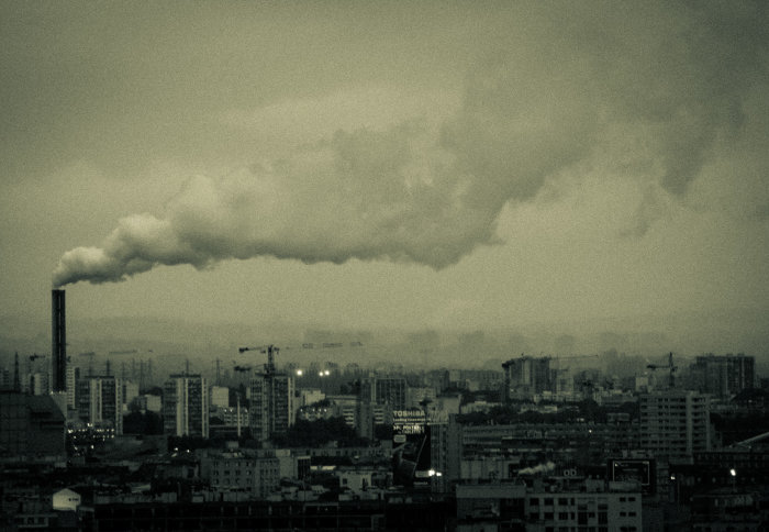 Smoke from a factory chimney belches out thick white smoke over a dark, foggy city-scape