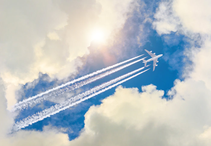 Aeroplane flies through a blue, cloudy sky - contrail trailing behind it.