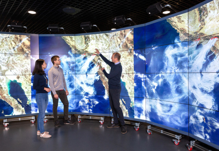 Professor Apostolos Voulgarakis standing in the Data Observatory, discussing with younger researchers. On the screens is displayed a satellite image of wildfires in California.