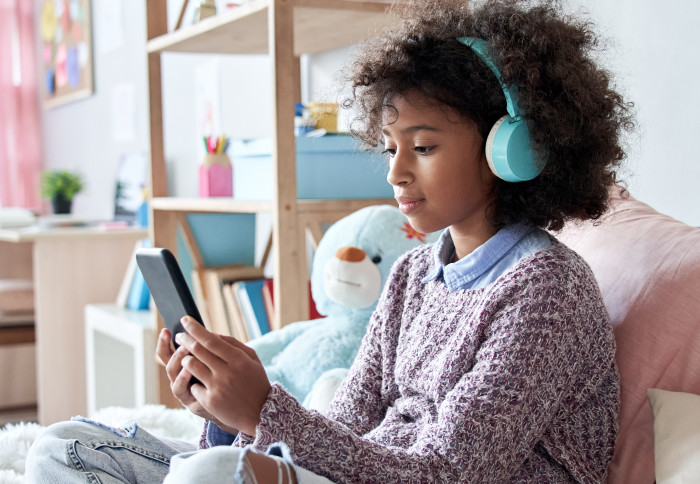 African-American girl wearing headphones holding phone sitting on bed.
