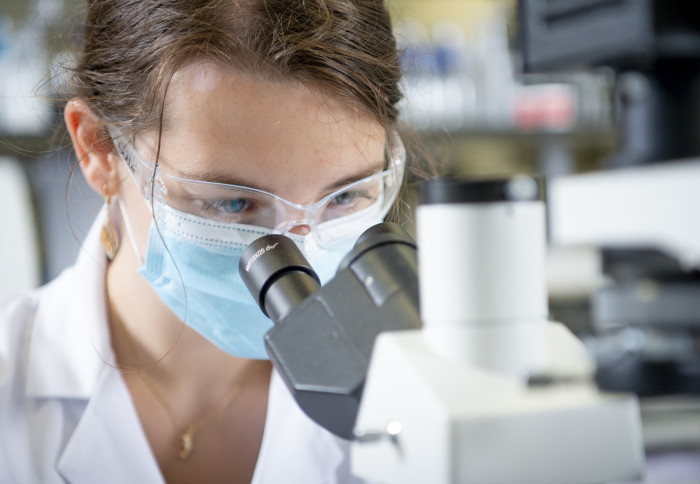 A member of the Multus Media team uses a microscope in the lab at the White City Incubator. She is wearing a mask and safety glasses and complying with COVID-19 safety regulations.