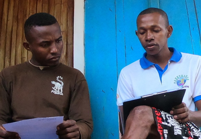 Photo shows a male researcher sitting next to another man while they fill out a survey on a clipboard. They are in Madagascar in a wooden-walled building, both men wear t-shirts.