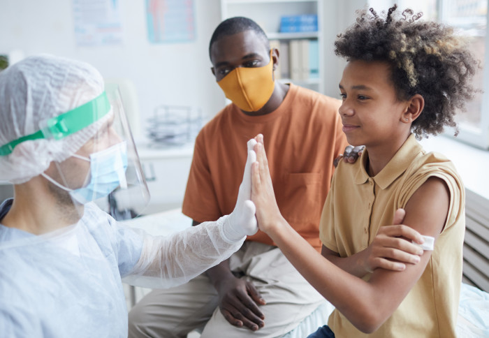 Child high fives doctor giving him his vaccination. His father looks on.