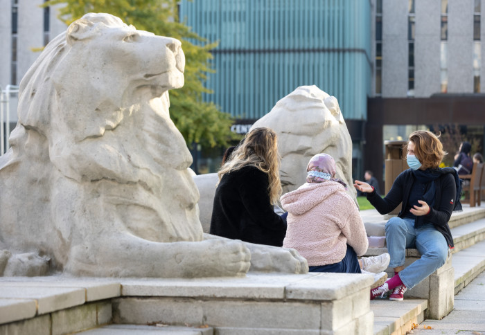 Students sat next to stone lions at Imperial. They are wearing face coverings and are chatting.