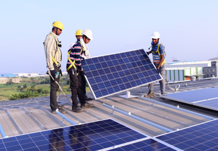 Photo shows a warehouse rooftop where four Indian men wearing hard-hats and safety harnesses are manoeuvring a solar panel into place among an array of others. The sky is blue and clear behind them.