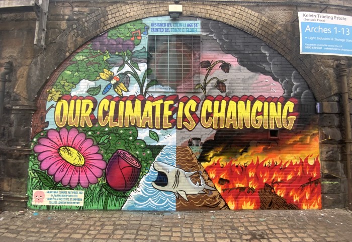 Photo of a street mural on a railway arch, reads "Our Climate Is Changing", one half is a flowery meadow, the other is a burning scorched earth scene