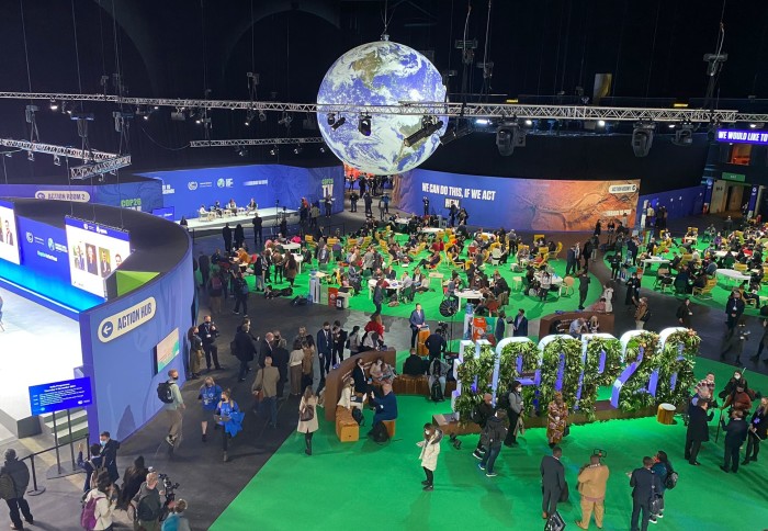 COP26 Venue space: people milling around in a conference venue looking at exhibits. A large sign reads COP26, a lit-up globe hangs from the ceiling