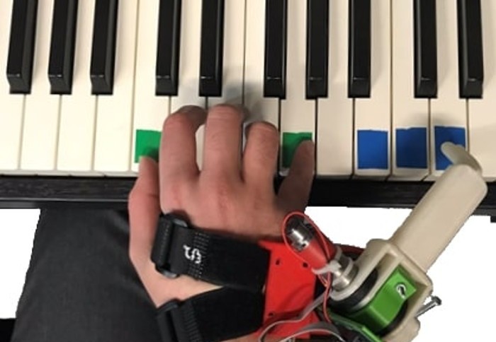 Close up photo of a hand equipped with an extra robotic thumb outside of the little finger. The robotic thumb is being used to play the piano.
