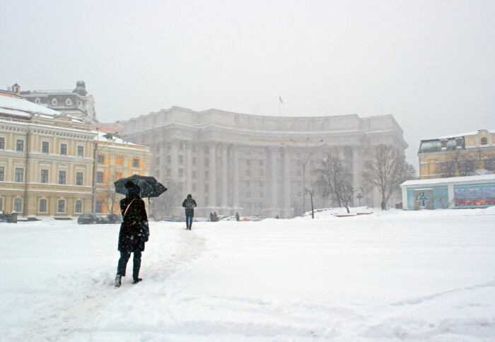 A person walks in the snow in Ukraine carrying an umbrella in front of old buildings.