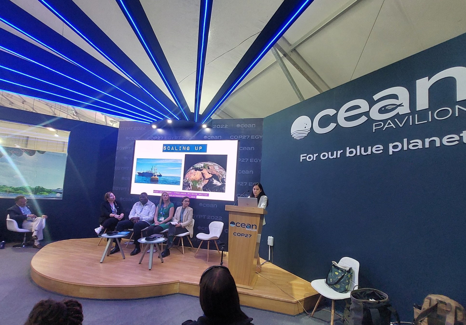 Photo shows a room with a stage, people are giving a presentation, the sign on the wall reads: Ocean Pavillion, For our blue planet
