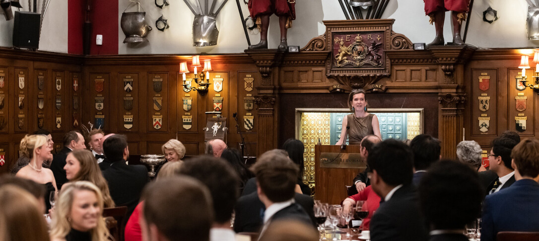 A snapshot from a previous dinner: a female speaker stands at a podium in a grand, old-fashioned hall in front of an audience. The dress code appears to be formal. 