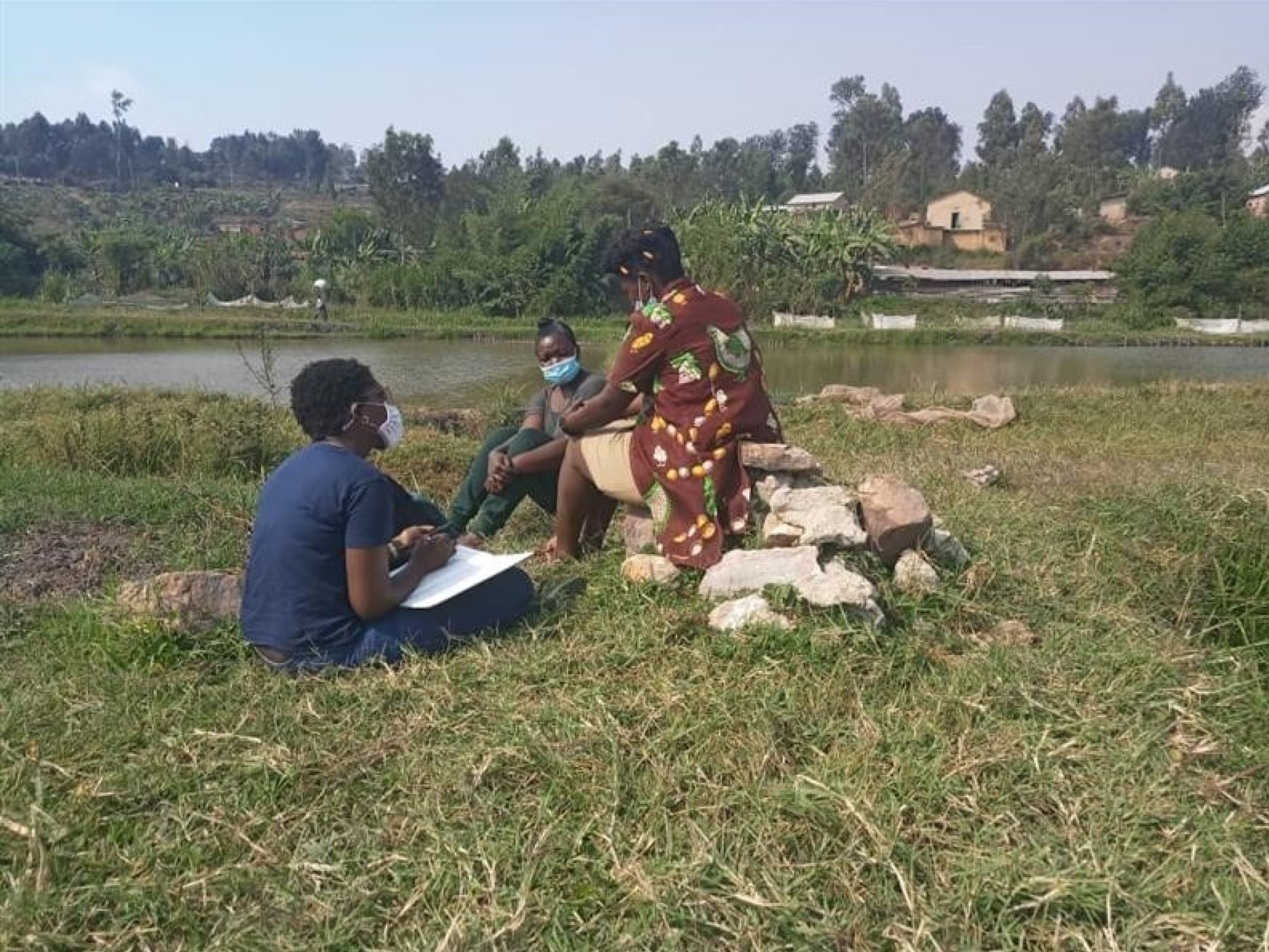 Three people sit outside by a river, wearing face masks 