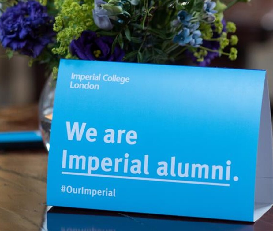 An Imperial branded tent card reading 'We are Imperial alumni #OurImperial' and showing the Imperial logo. The tent card is placed on a table with a backdrop of flowers, indicating the photo was taken at an event