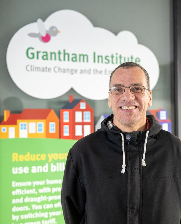 Ralf Toumi, a white man in his fifties wears a dark jacket with white hood drawstrings, he has dark short hair and glasses. He stand in front of a colourful full height window-sticker that reads Grantham Institute in a cloud shape.