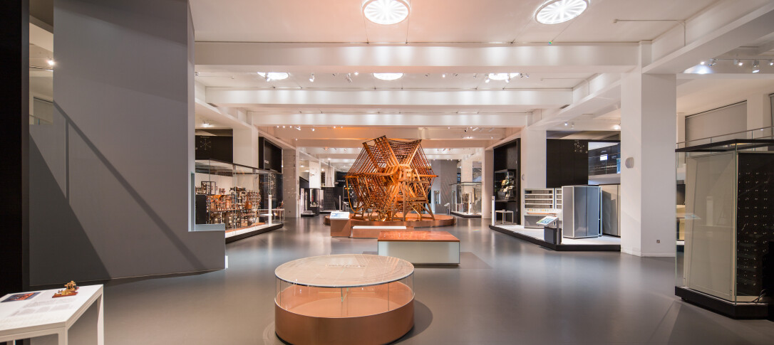 A landscape photo of The Information Age Gallery at the Science Museum, it appears to be spacious, contemporary in its lighting and devoid of people for the photo. Pictured in the centre background is a high copper and wood aerial tuning inductor from Rugby Radio Station. 