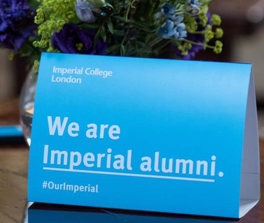 An Imperial branded tent card reading 'We are Imperial alumni #OurImperial' and showing the Imperial logo. The tent card is placed on a table with a backdrop of flowers, indicating the photo was taken at an event