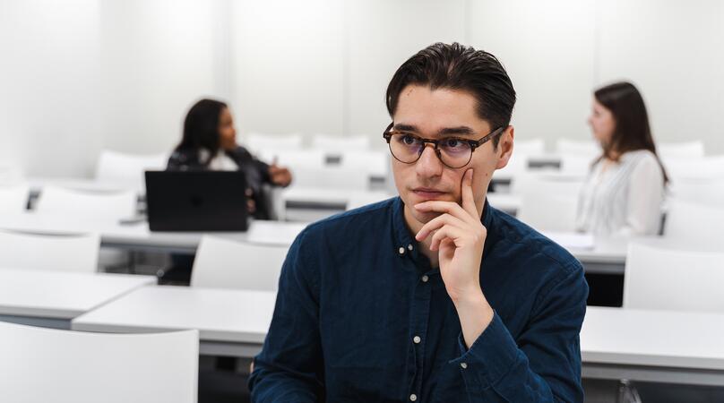 A young male student with glasses sitting in a classroom with two classmates behind in the distance