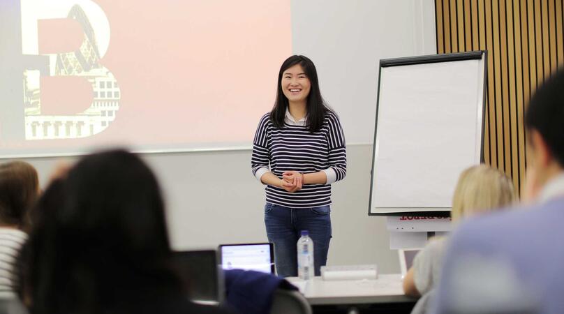 A women in front of a white board with the red IB logo on it speaking to students