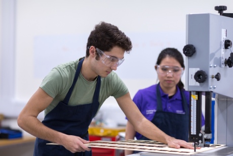 A student using a bandsaw to cut a sheet of thin wood, with a student mentor watching