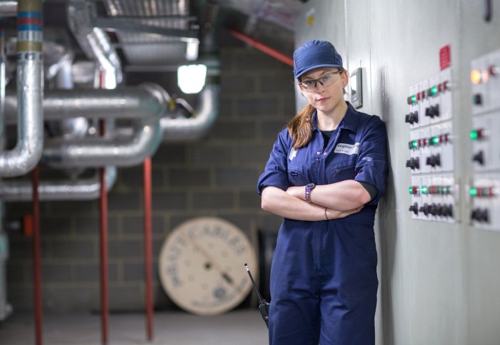 young woman in dark blue overalls with metal pipes and coloured switches in background