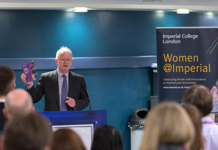 man standing at lectern holding leaflet, with Women@Imperial banner in background