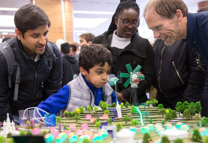 People crowded round a table top model city with wind turbines, solar panels and light-up houses