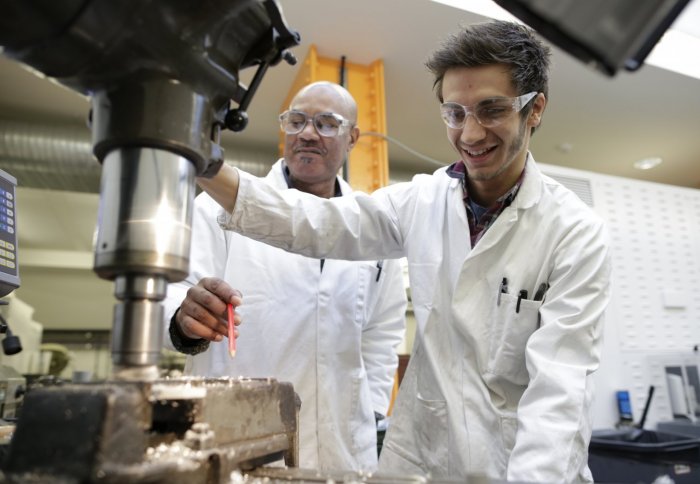 two men in lab coats and safety goggles, one using a piece of machinery
