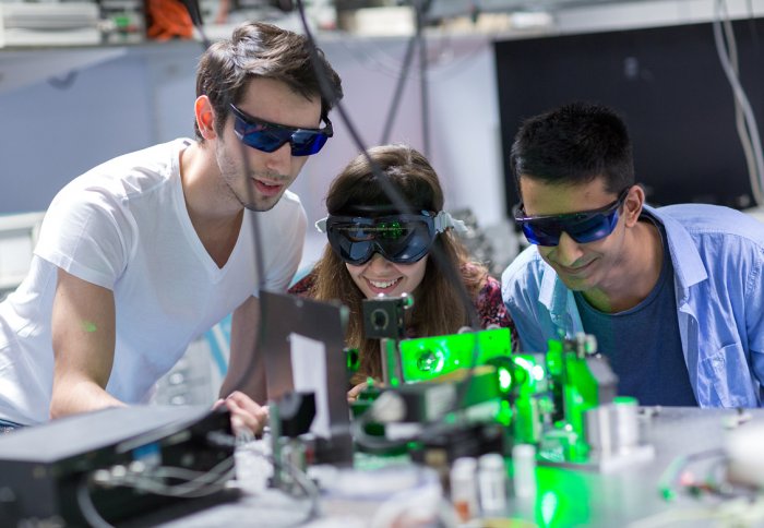 Imperial students in a lab wear protective goggles while looking into a microscope