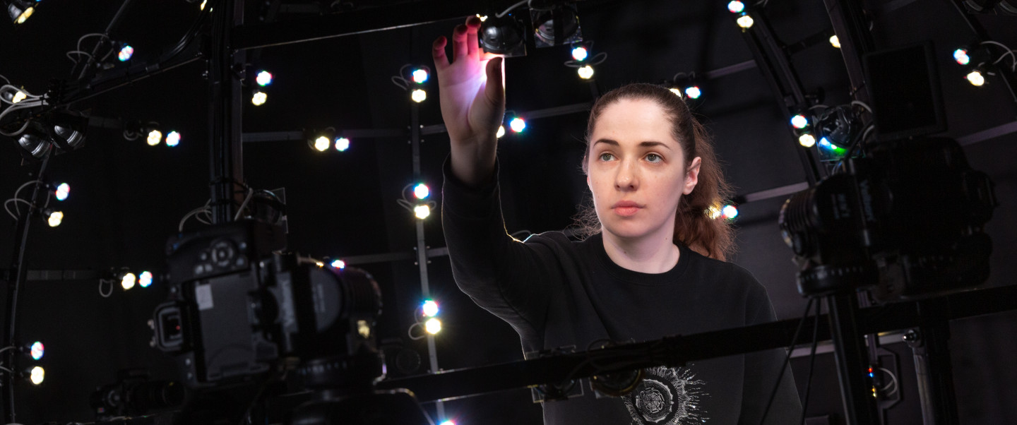 Light-skinned woman wearing a black t-shirt touches a light above her face. The woman is surrounded by other lights.