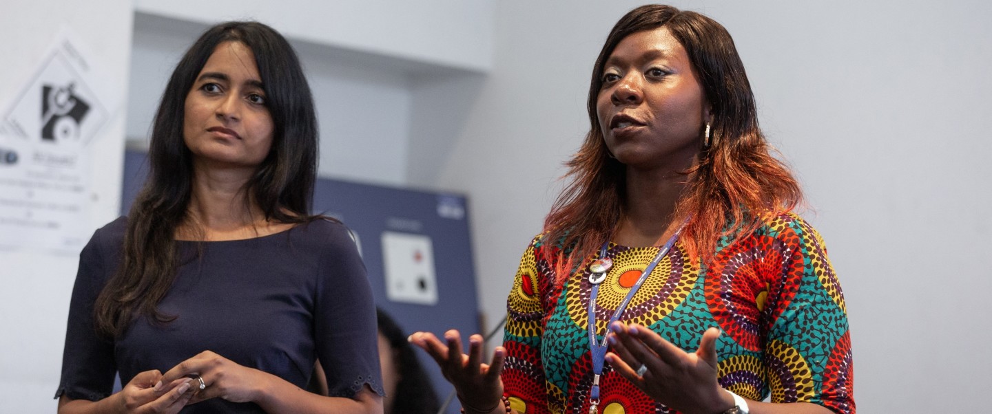 Two women talk to audience in a lecture theatre. They are shown gesticulating with their hands.