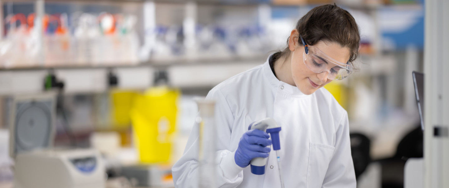 Light-skinned woman wearing goggles, blue gloves and white lab coat stands in laboratory setting